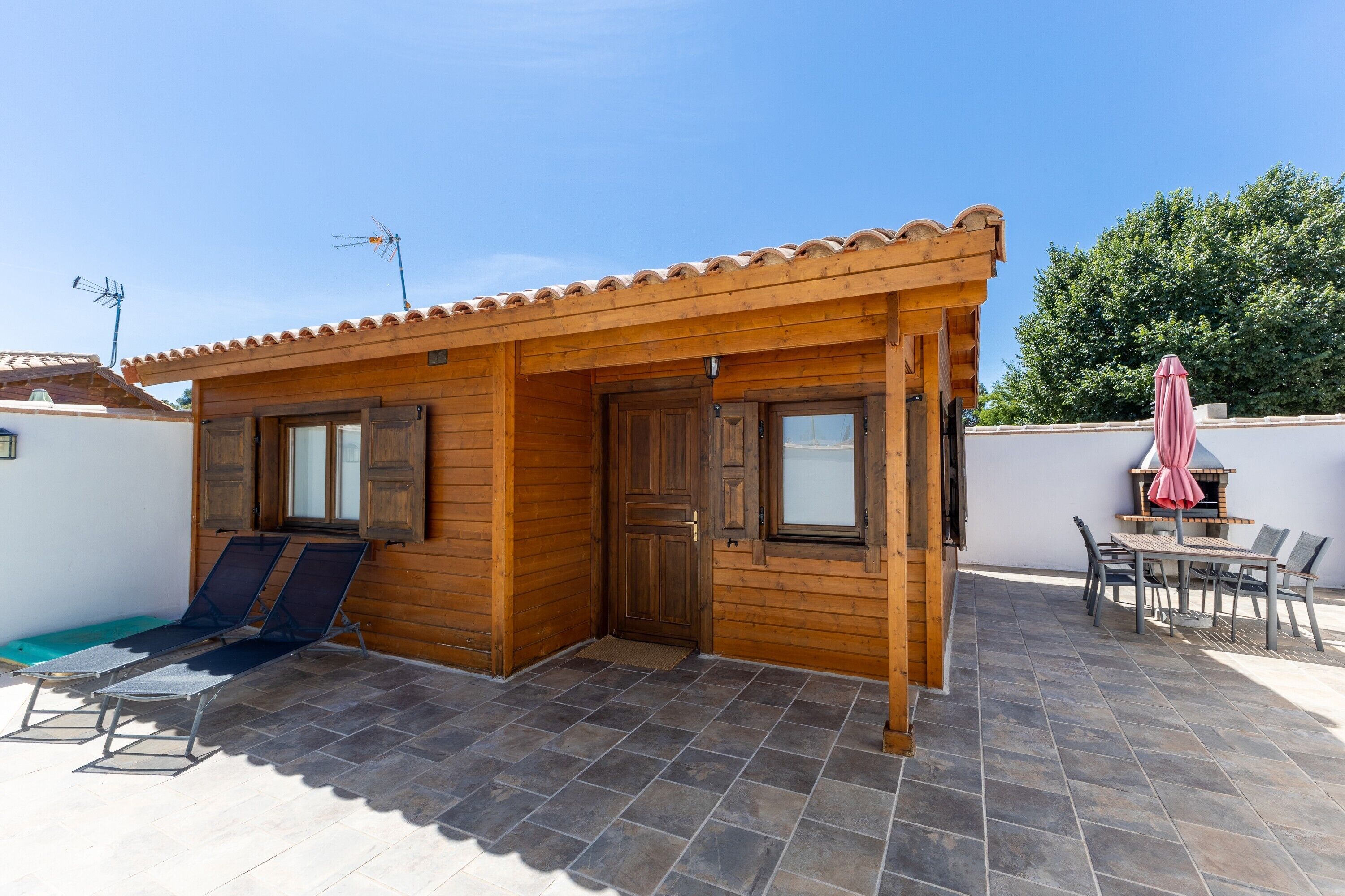 En træhytte med fliseterrasse, swimmingpool og et bord med stole.