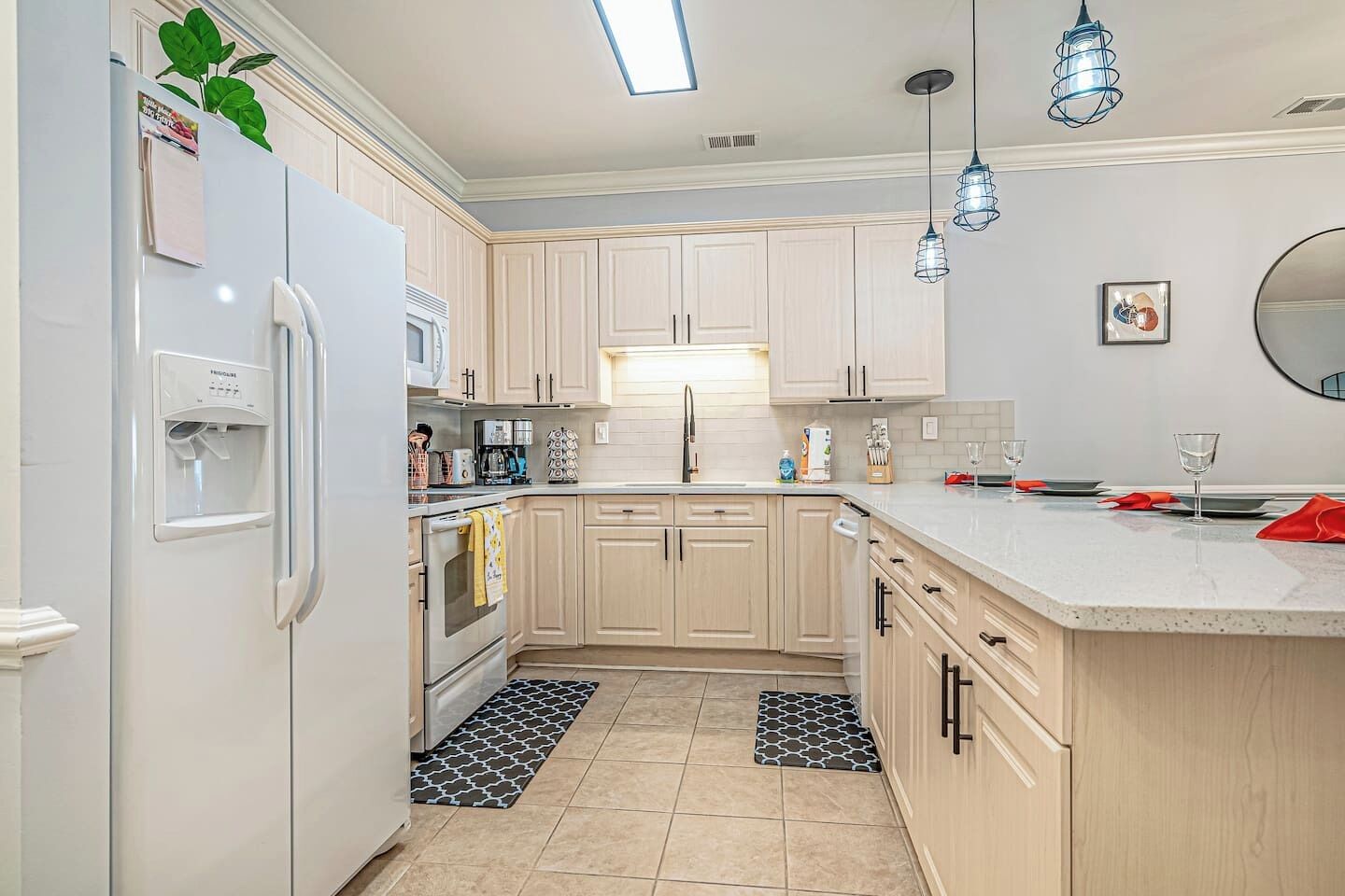 A modern kitchen with light wooden cabinets, a white refrigerator, a built-in oven, a microwave, and a sink. There are hanging pendant lights and a round mirror on the wall.