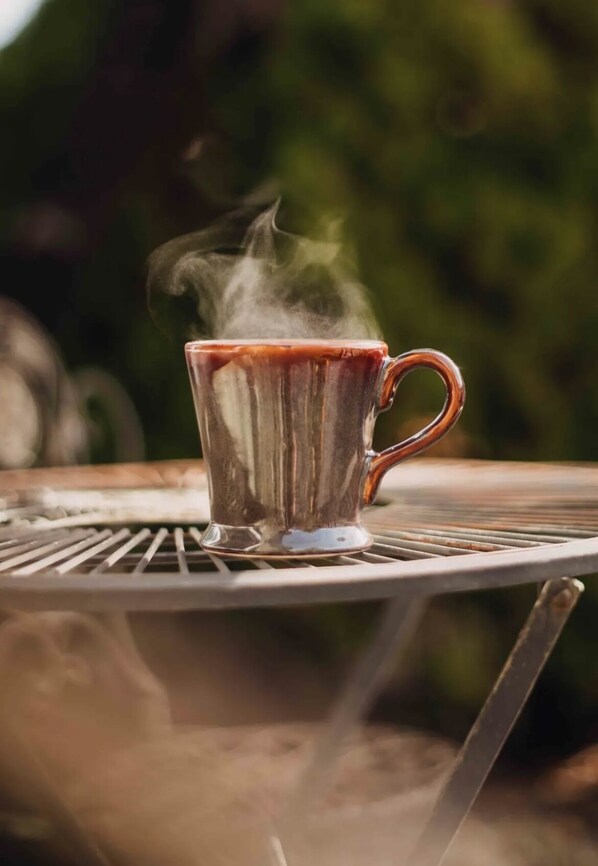 A clear glass mug with steam rising from it, placed on a metal table outdoors.