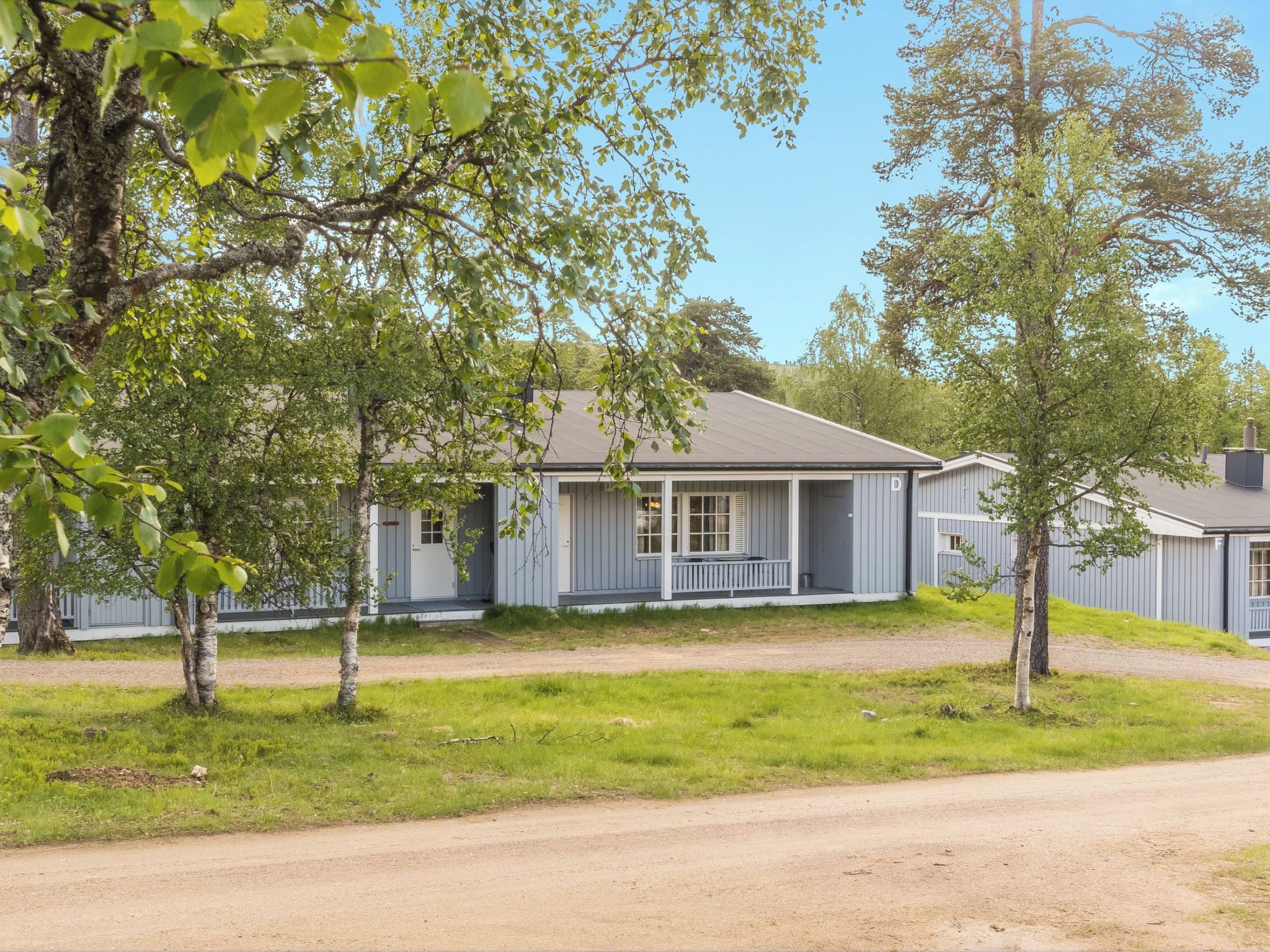 Eenlaags huis met veranda, omringd door bomen en een onverharde weg.