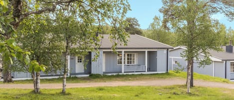 Eenlaags huis met veranda, omringd door bomen en een onverharde weg.