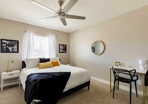 Quarto com cama, escrivaninha, cadeira, ventilador de teto e duas imagens emolduradas na parede.