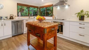 A modern kitchen with white cabinetry, a wooden table with a fruit bowl, and a view of greenery through large windows. - Peaceful Lake Rotoiti Escape | Jetty, Kayaks & Clear Water Views (Rotorua)