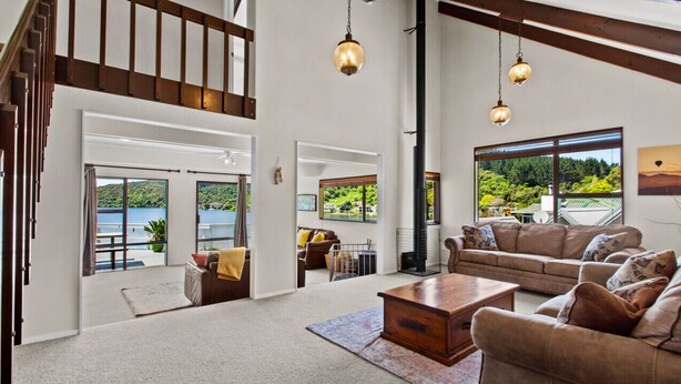 A spacious living room with a large sofa, a coffee table, and a fireplace. There are large windows offering a view of the outdoors. - Peaceful Lake Rotoiti Escape | Jetty, Kayaks & Clear Water Views (Rotorua)