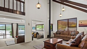 A spacious living room with a large sofa, a coffee table, and a view of a scenic landscape through large windows. - Peaceful Lake Rotoiti Escape | Jetty, Kayaks & Clear Water Views (Rotorua)