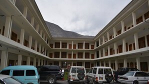 Un immeuble à plusieurs étages doté d’une cour intérieure. Plusieurs véhicules sont stationnés dans la cour, notamment un fourgon bleu, un VUS noir et un VUS blanc. L’immeuble possède de nombreux balcons avec des garde-corps en bois.