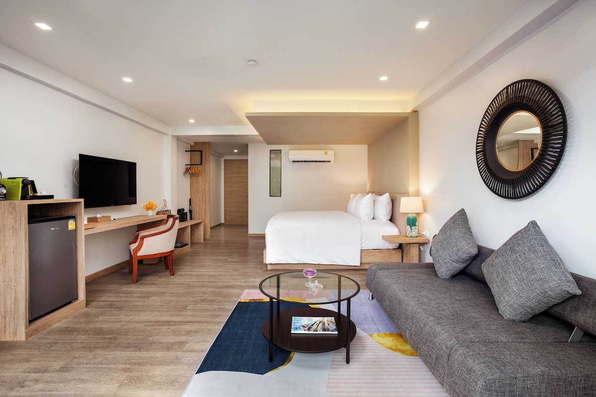 A modern hotel room with a bed, a sofa, a desk, a TV, and a small fridge.