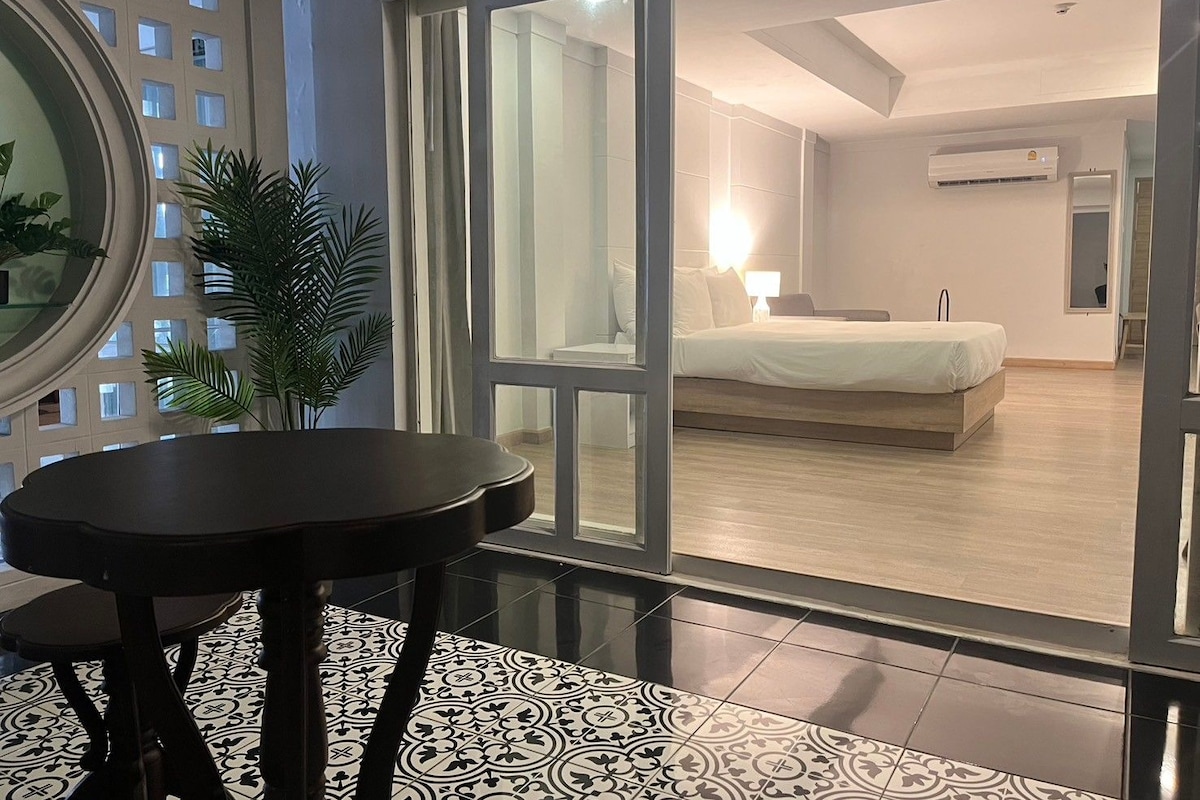 Una habitación de hotel moderna con un amplio ventanal corredero que da a un balcón, una cama con cabecera blanca, una mesita de noche y una planta en maceta.