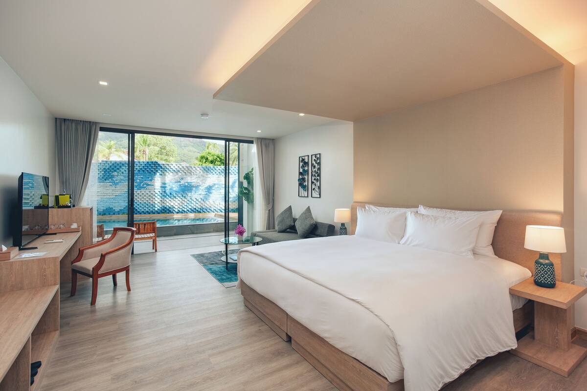 A modern hotel room with a large bed, a desk, and a chair. There is a view of a pool and greenery outside the window.