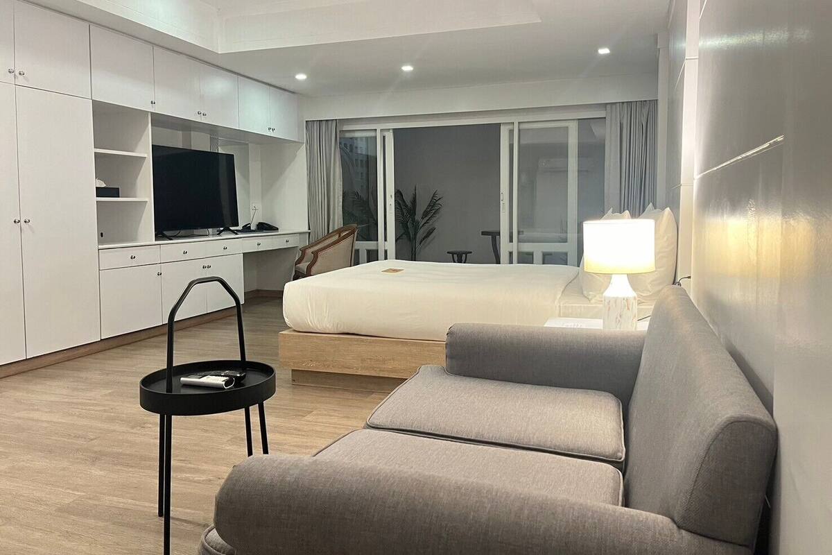 A modern hotel room with a bed, a sofa, a desk with a TV, and a window with curtains.