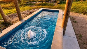 A rectangular swimming pool with a water feature in the center, surrounded by a concrete deck, set against a sunset sky with a grassy landscape. - Beachfront | Pool | Ocean Views, Loft & Bar (Gilchrist)