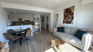 A modern living room with a grey sofa, a dining table with wicker chairs, a kitchen area with a fridge and microwave, and a large painting on the wall. - Perla del Mar - one step from the beach and center of Platja d'Aro (Castell-Platja d'Aro)