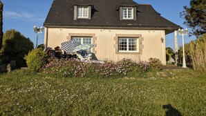 Een tweelaags huis met een donker dak, beige muren en een klein voortuintje. Er zijn twee ramen aan de voorkant en een schoorsteen.