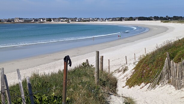 Een strand met een houten hek, een heldere lucht en huizen op de achtergrond.