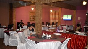A banquet hall with round tables covered in white tablecloths and red drapes, chairs with white covers, and a television mounted on the wall. - Gilgit Gateway Hotel (Gilgit)