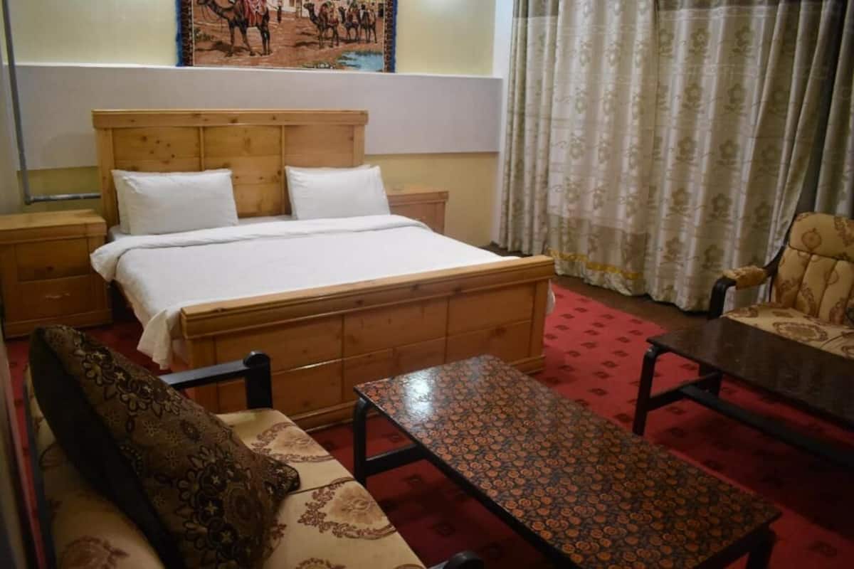 Habitación de hotel con una cama de madera, una mesita de noche, un sofá estampado, una pequeña mesa y un sillón con estampado.