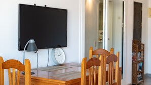 Un coin repas comprenant une table en bois, quatre chaises, un téléviseur fixé au mur, une lampe de bureau et une horloge.