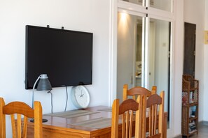 Ruokailutila, jossa on puinen pöytä, neljä tuolia, seinälle asennettu televisio, pöytävalaisin ja kello.