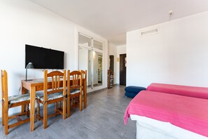 A room with a TV, wooden dining table and chairs, a bed with pink bedding, and a sliding door leading to another room.