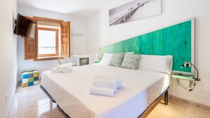 Une chambre d’hôtel avec un lit, un téléviseur fixé au mur, une fenêtre avec des volets en bois et un tableau au-dessus du lit.