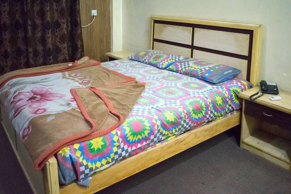 A bed with a colorful quilt, a brown blanket, and a floral throw. There is a bedside table with a phone and a lamp.