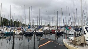 Uma marina com vários barcos atracados, um helicóptero no céu e um céu nublado.