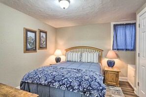 Un dormitorio con cama, dos lámparas de noche, una mesita de noche de madera, una ventana con cortinas y dos cuadros en la pared.