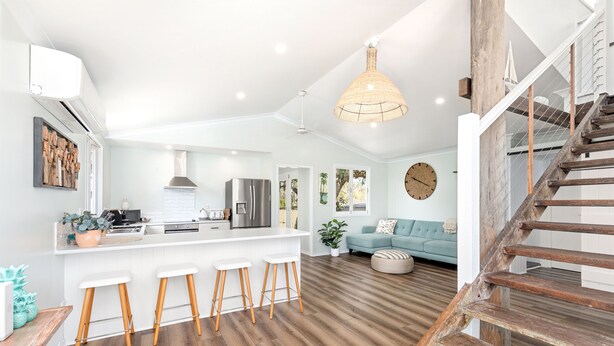 A spacious kitchen with a central island, a dining area with a blue sofa, and a wooden staircase. - Villa Sunshine - Pet Friendly Fully Air conditioned (Toogoom)