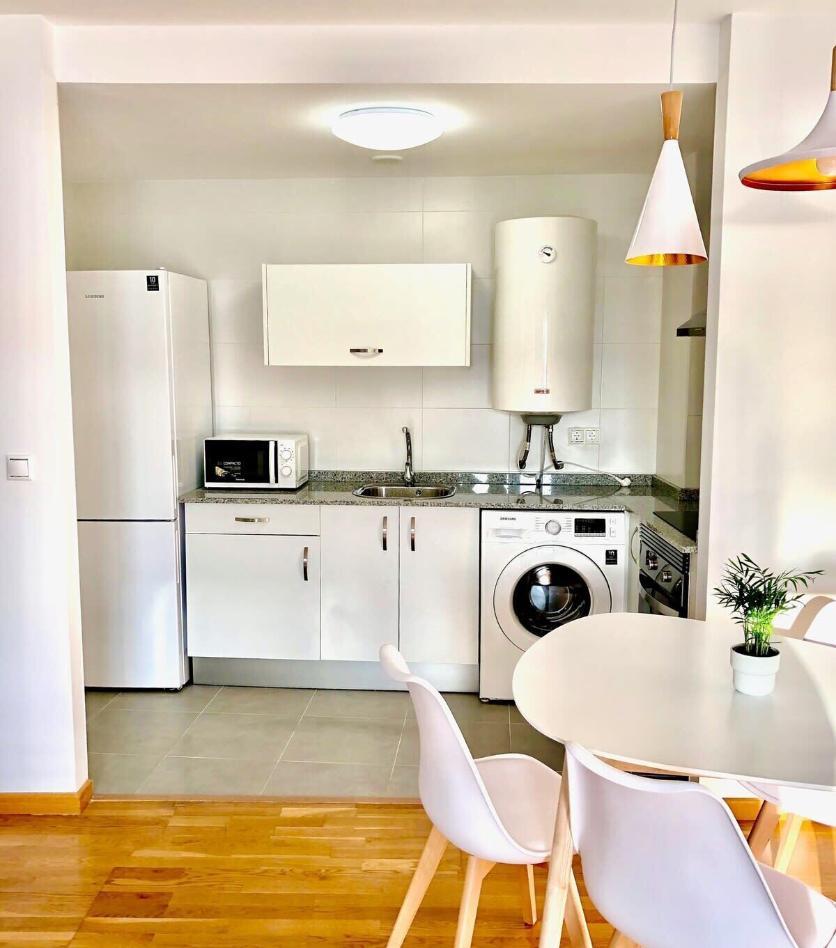 A modern kitchen with white cabinets, a built-in microwave, a washing machine, and a dining area with a white table and chairs.