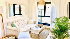 A living room with a white sofa, a coffee table, a large window with curtains, a hanging light fixture, and a potted plant. - The Cliffs - Sunset Village Beach 3E (Viveiro)