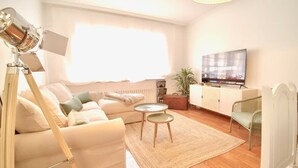 A well-lit living room with a sofa, two side tables, a television, and a large window. - The Cliffs - Le nid de Martina (Porto do Barqueiro)