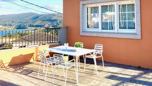 A patio with a table and chairs, a window with a view of mountains, and a balcony railing. - The Cliffs - Le nid de Martina (Porto do Barqueiro)