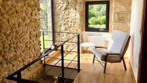 A room with stone walls, a wooden floor, a white chair, a small table, and a window with a view of greenery. - The Cliffs - Galleon Amieiro (O Vicedo)
