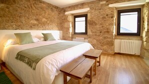 A bedroom with a stone wall, a bed with white bedding, two windows, a radiator, and a wooden bench. - The Cliffs - Galleon Amieiro (O Vicedo)