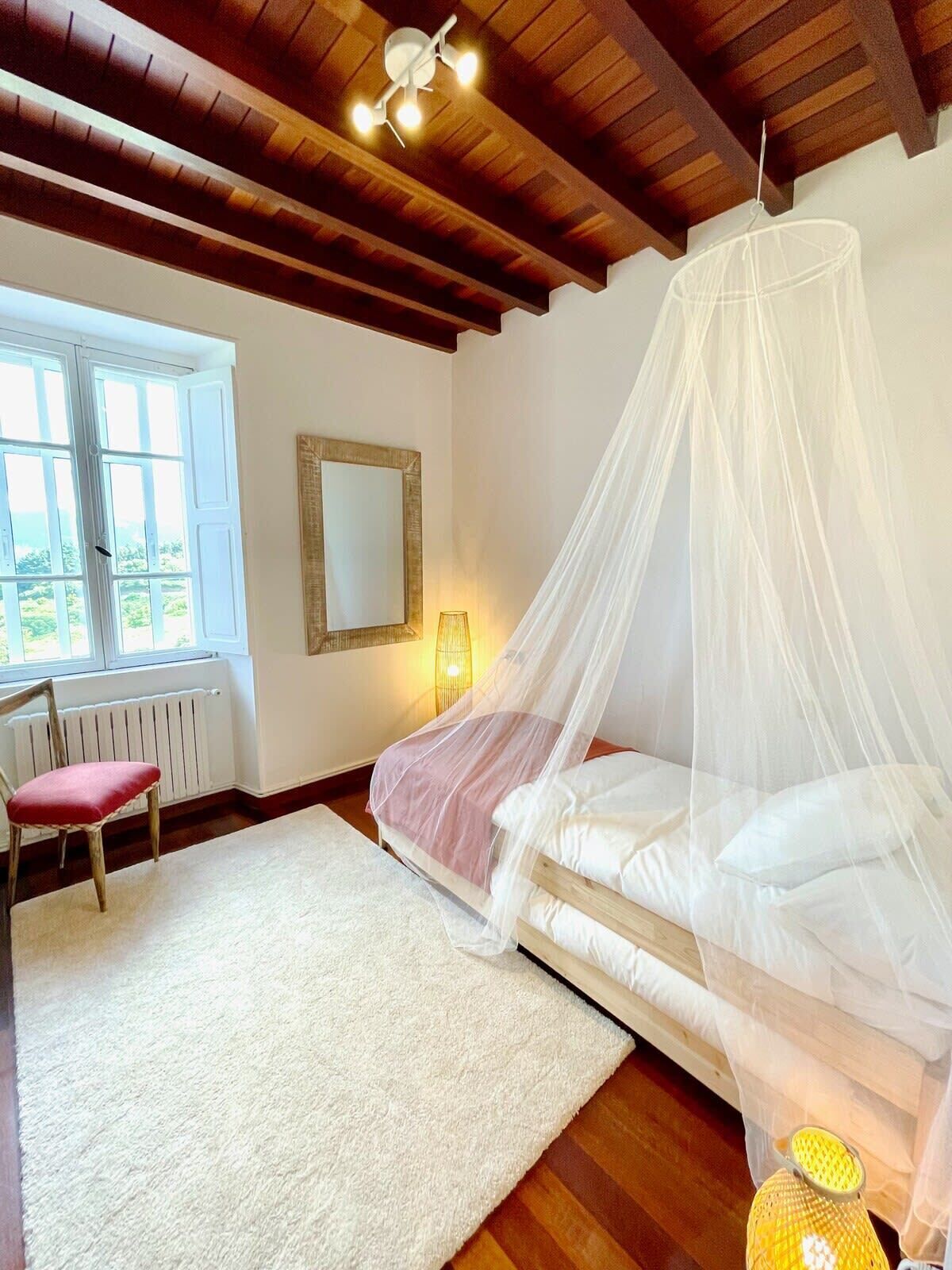 A bedroom with a bed under a canopy, a chair, a lamp, and a window with a view.