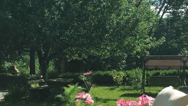 Un área de estar al aire libre con mobiliario de mimbre, que incluye un banco acolchado, rodeada por un jardín con flores rosadas y césped verde.