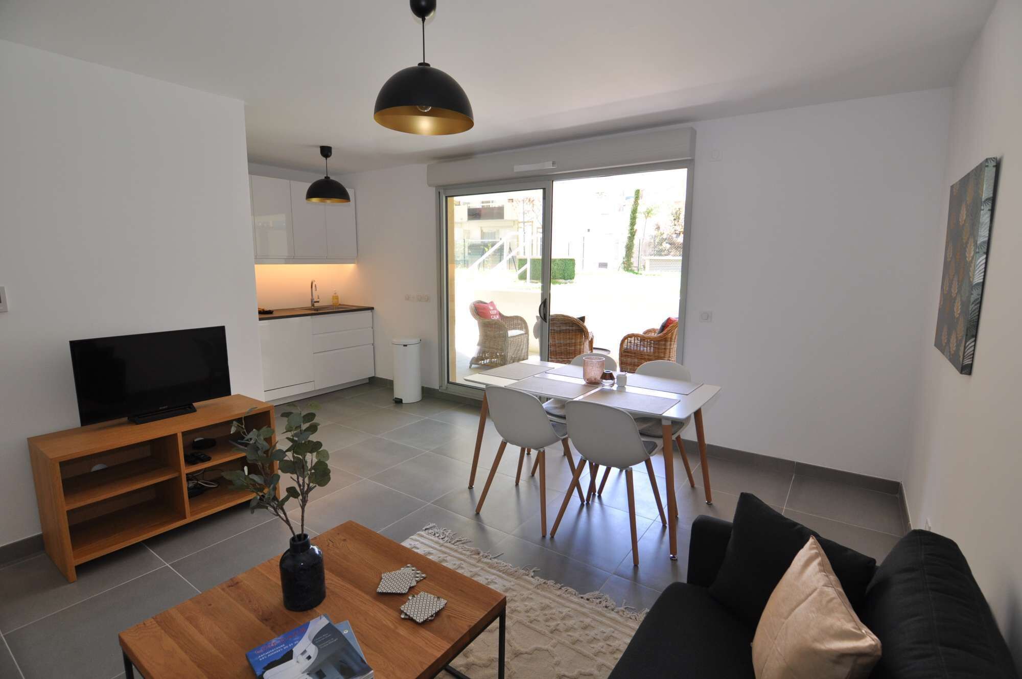 Un soggiorno moderno con una TV a schermo piatto, un tavolino in legno e un divano nero. C'è una zona pranzo con tavolo e sedie bianche, e una cucina con mobili bianchi e una lampada a sospensione nera.