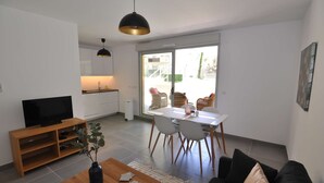 A modern living room with a flat-screen TV, wooden coffee table, and a black sofa. There's a dining area with a white table and chairs, and a kitchen with white cabinets and a black pendant light.