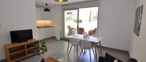 A modern living room with a flat-screen TV, wooden coffee table, and a black sofa. There's a dining area with a white table and chairs, and a kitchen with white cabinets and a black pendant light.