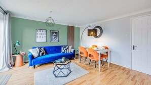 A modern living room with a blue sofa, orange chairs, a dining table, and a geometric pendant light.