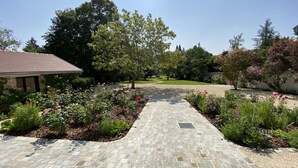A paved pathway leading to a house with a stone facade, surrounded by a well-maintained garden with various plants and flowers. - Charming Renovated Cottage in 2-Hectare Park with Fishing Pond, Near Forest, Close to Versailles (Senlisse)