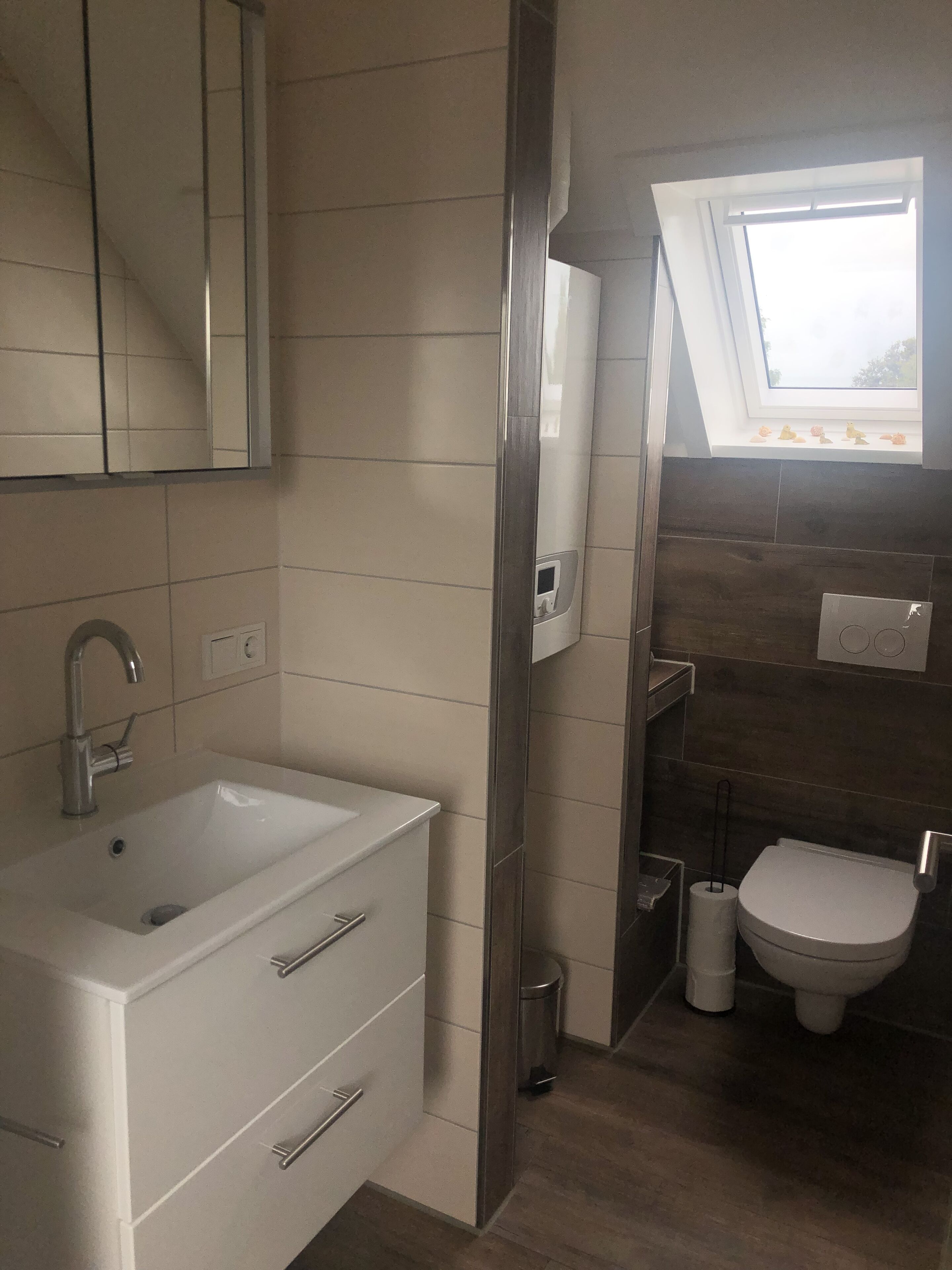 Et moderne badeværelse med en hvid vask, et spejl og et toilet. Der er et vindue over toilettet, og en lille hylde med pynt.