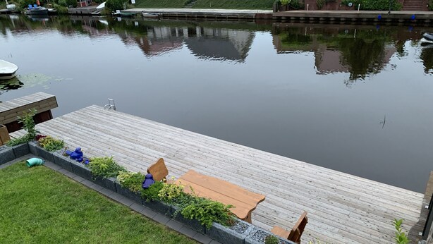 Una terraza de madera con sillas y una mesa, rodeada por un jardín con plantas y césped.