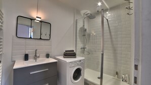 A modern bathroom with a built-in washing machine, a glass shower enclosure, and a vanity with a mirror and sink. - Magnificent T2 hyper center, hyper equipped (La Rochelle)