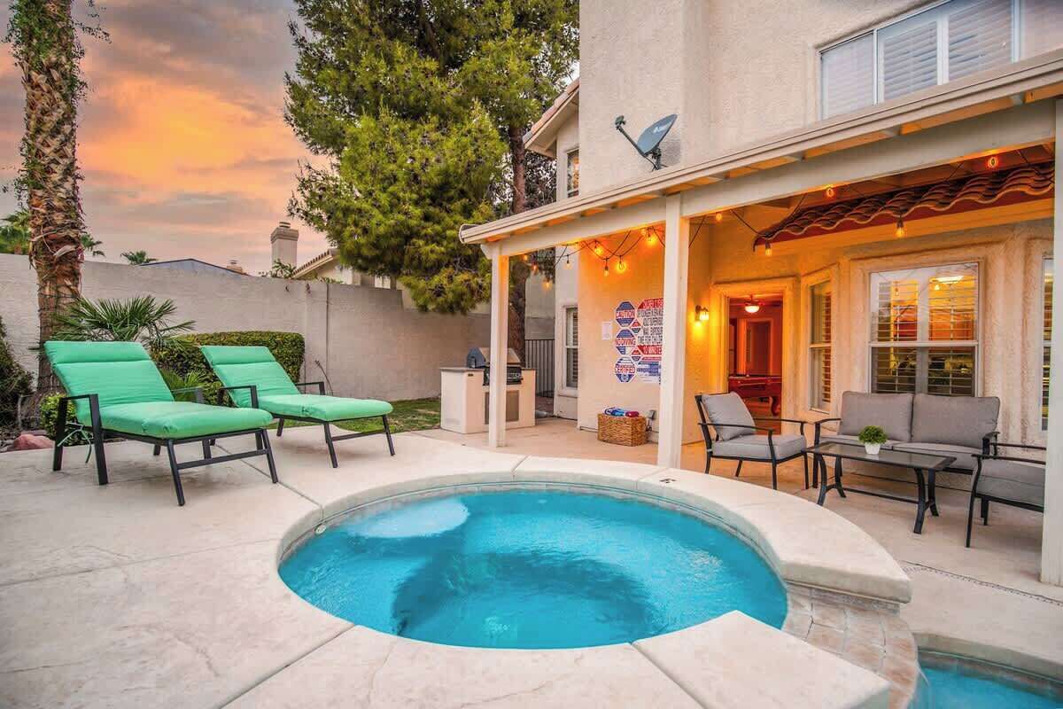Un jardin avec une piscine, des fauteuils de jardin verts et un patio couvert aménagé avec du mobilier d’extérieur.