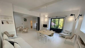 Un salón moderno con un sofá blanco, una mesa de comedor con sillas, sillas de mimbre y un ventanal con vistas a zonas verdes.