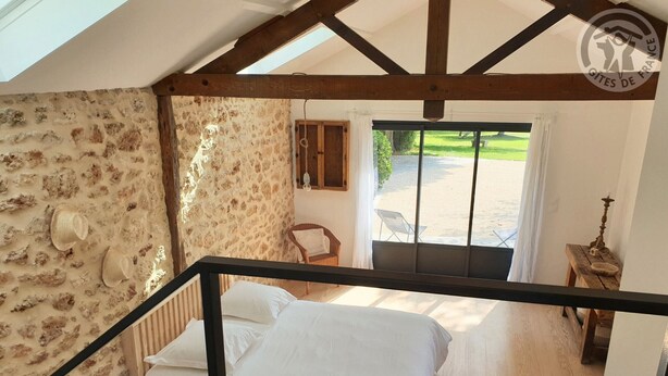 Un dormitorio con cama, una silla de madera, una mesita y vista a un jardín por la ventana.