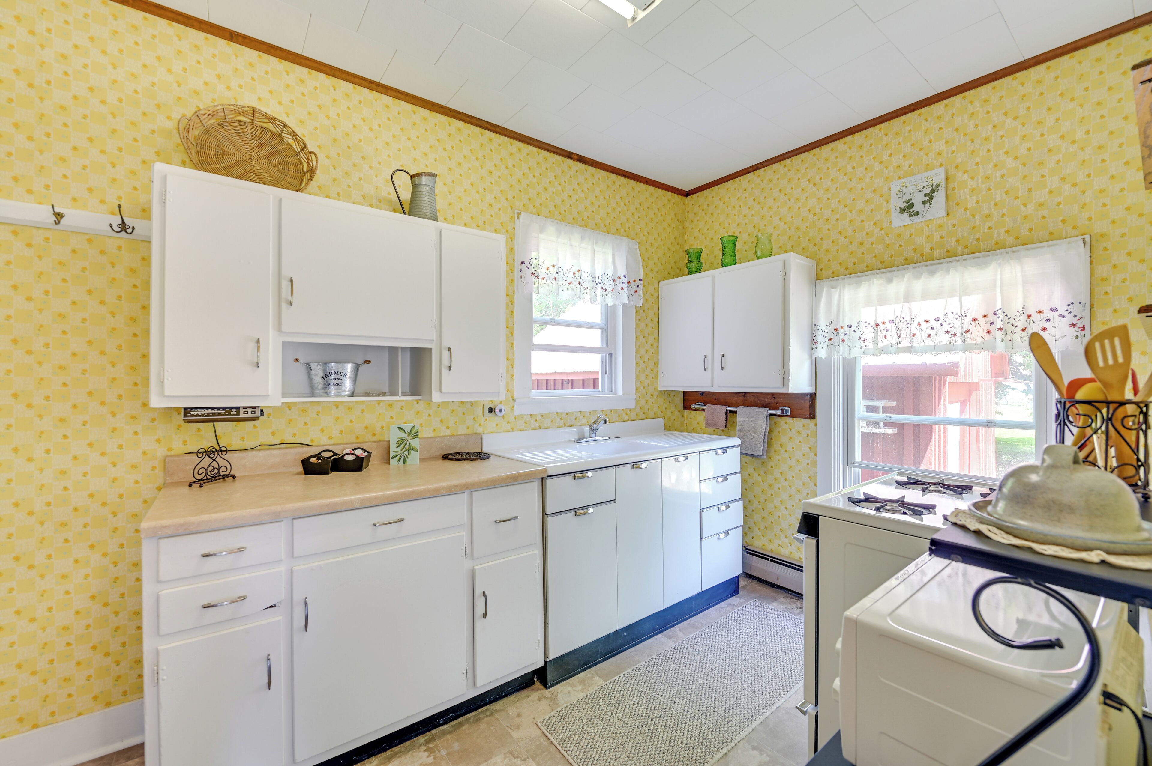 A kitchen with white cabinets, a sink, and a stove. There is a window with a floral curtain and a basket on the wall.