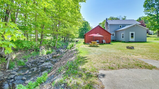 A red barn with a picnic table in a grassy yard, a gray house, and a stream with rocks. - Countryside Hideaway - 10 Mi to Cooperstown! (Westford)
