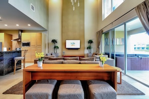A modern living room with a wooden dining table, grey sofas, and a flat-screen TV mounted on the wall.